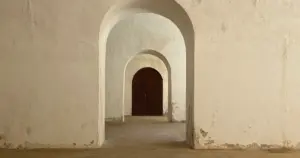 A series of arched stone doorways leading to a closed wooden door, lit by soft natural light.